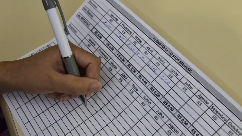 Mão anota valores em reais em uma planilha impressa de gastos mensais.