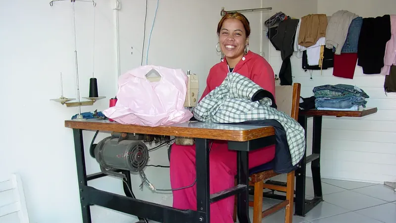 Costureira sorri em seu ateliê em São Francisco do Sul, diante de tecidos e de uma máquina de costura.