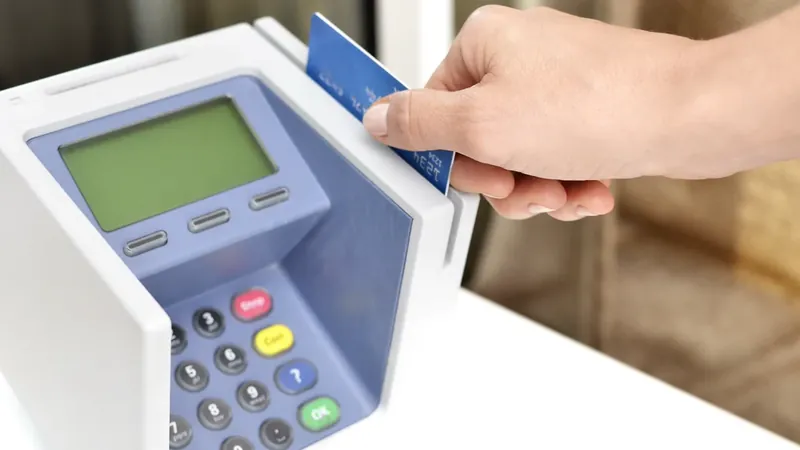 Mão passa um cartão azul na lateral de uma máquina de pagamento sobre um balcão branco.