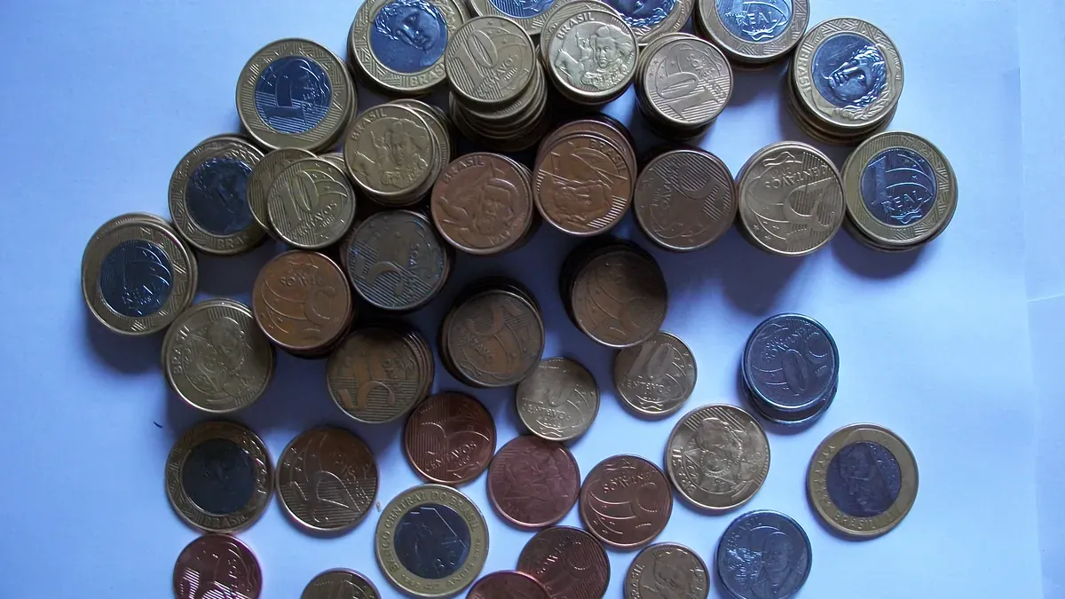 Moedas brasileiras de real e centavos formam pequenas pilhas sobre uma superfície azul-clara.
