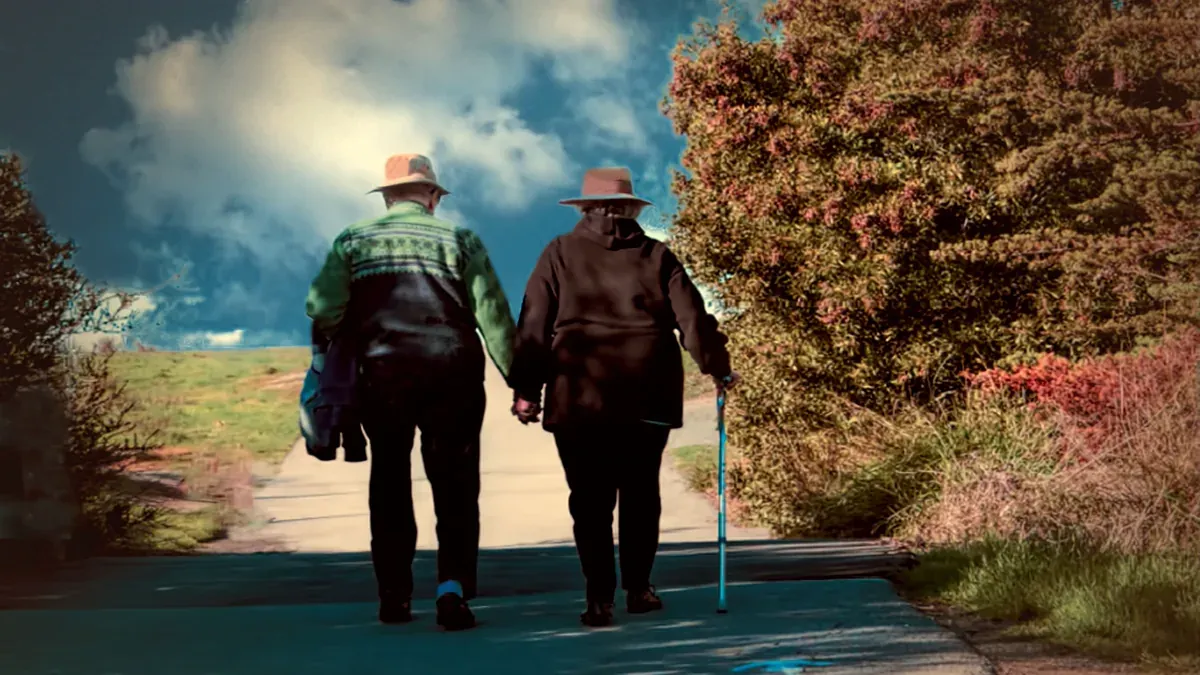 Two older adults walking hand in hand along a tree-lined path beneath a blue sky.